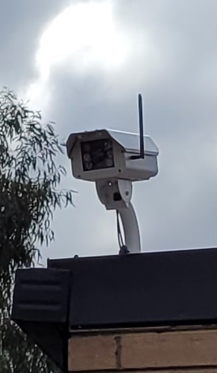 A white sensor, which looks like a security camera, on top of a shed with a cloudy sky in the background