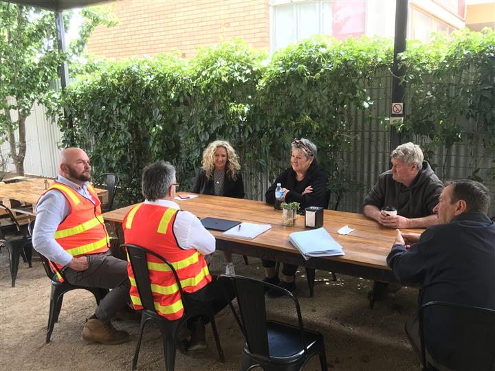 A group of people sitting outside around a table. 2 men are wearing hivis vests, and there are 2 women and two men around the table. 