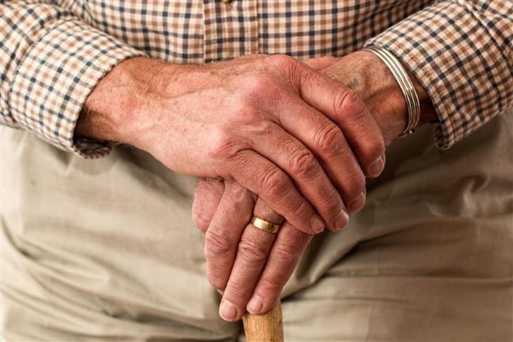 Elderly person with hands on their walking stick. They are wearing a checkered shirt and tan pants. They have a gold wedding ring on their right index finger. 