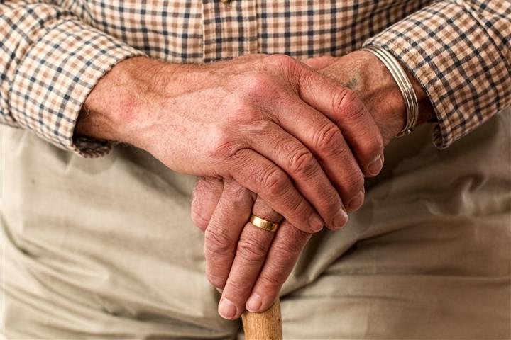 Elderly person with hands on their walking stick. They are wearing a checkered shirt and tan pants. They have a gold wedding ring on their right index finger.