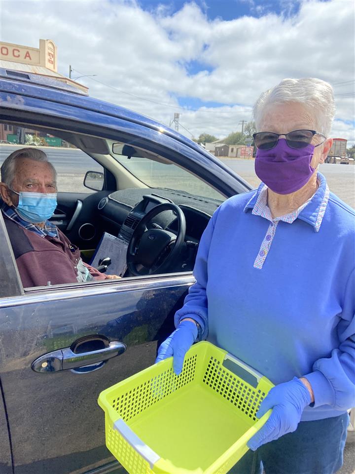 Two meals on wheels volunteers. One is sitting behind the drivers seat of a blue car on the left and the other person is standing outside the car, holding a bright green container. Both are wearing blue facemasks. 