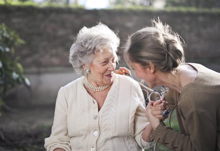 Two women sitting side by side laughing. One woman is elderly, with a cardigan on and a pearl necklace on, the other is younger with a brown top on and her face is turned towards the elderly lady. 